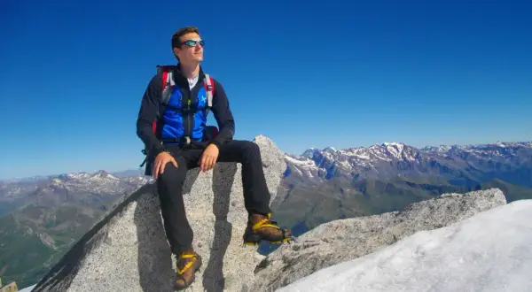 Giovane alpinista seduto sulla cima Presanella, ammira il panorama mozzafiato delle Alpi sotto un cielo azzurro.