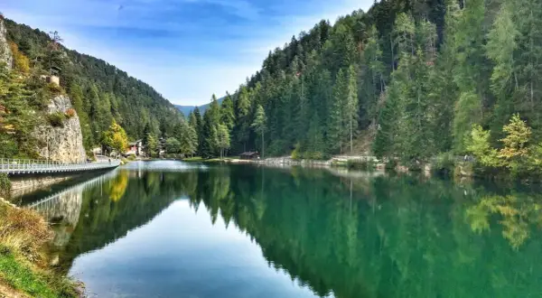 Un **lago smeraldo** incastonato tra montagne boscose, con un ponte pedonale che riflette le sue acque cristalline.