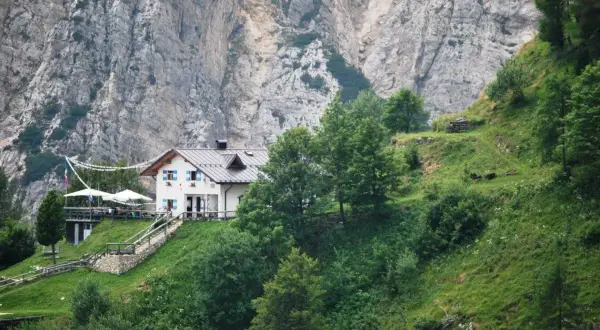 Rifugio Casarota, un'accogliente struttura bianca con persiane blu, incastonata nel verde di un pendio montano, sotto imponenti pareti rocciose.