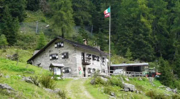 Il rifugio Trivena, con bandiera italiana, è una casa in pietra immersa nel verde, con un trattore parcheggiato accanto.