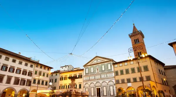 Piazza di Empoli illuminata da luci natalizie, con la fontana, la chiesa e il campanile che si stagliano sotto un cielo azzurro.