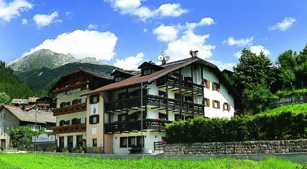 Albergo La Montanara a Predazzo, con le sue balconate in legno e le montagne sullo sfondo, incorniciato da un cielo azzurro e nuvole bianche.