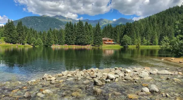 Il lago dei caprioli, circondato da fitti boschi e montagne, riflette un cielo azzurro con nuvole bianche. Una casa in legno si affaccia sull'acqua.