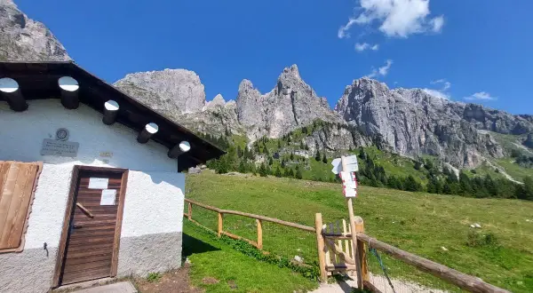 Malga Cavallera: sentiero con cartelli direzionali e rifugio bianco ai piedi delle maestose montagne.