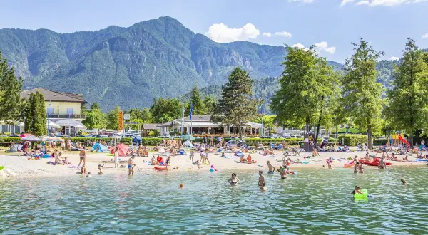 Spiaggia affollata al camping Riviera Caldonazzo, con gente che prende il sole e nuota nel lago, montagne sullo sfondo.