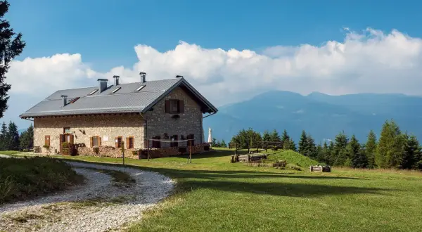 Malga Campo, un rifugio in pietra con tetto metallico, immerso nel verde di un prato alpino, con montagne sullo sfondo.