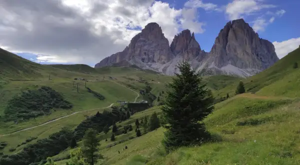 Verdi pendii alpini verso le imponenti cime del Sassolungo, con sentieri e impianti di risalita. Immagina le cascate basaltiche di Canazei in lontananza.
