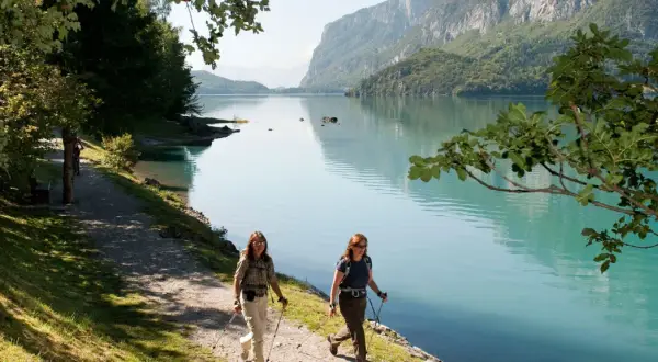 Due escursioniste camminano lungo il sentiero del lago di Molveno in estate, con montagne verdi sullo sfondo.
