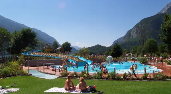 Giornata di sole all'acquapark Molveno, con scivoli blu, piscine piene di gente e montagne sullo sfondo.