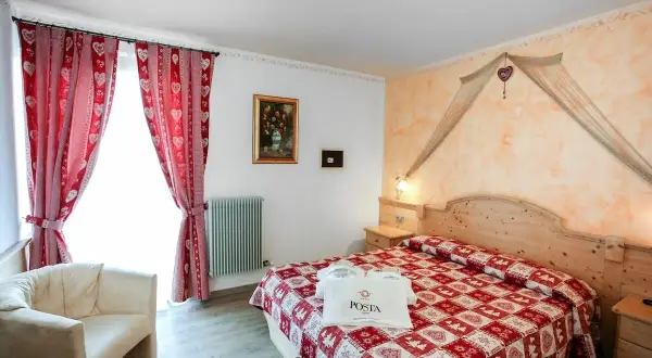 Camera accogliente con letto a quadri rossi, tende a cuoricini e un sacchetto con logo POSTA HOTEL 1899.