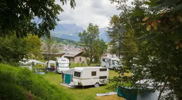 Un tranquillo camping calavise con roulotte e camper immersi nel verde, sullo sfondo un villaggio alpino e montagne nebbiose.