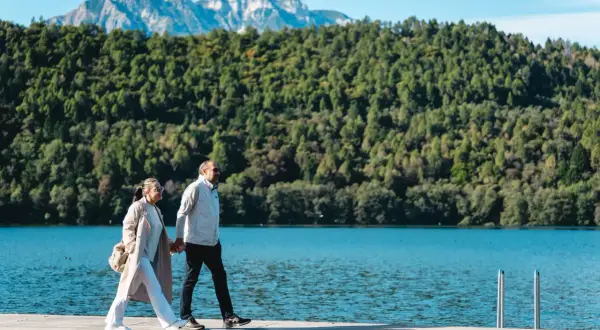 Coppia passeggia mano nella mano sul sentiero dei pescatori a Levico, con lago e montagne sullo sfondo.