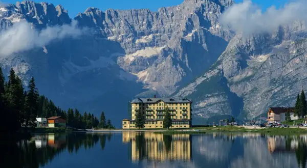 Un hotel giallo si specchia in un lago alpino, con imponenti montagne sullo sfondo: ecco dove si trovano le Dolomiti.