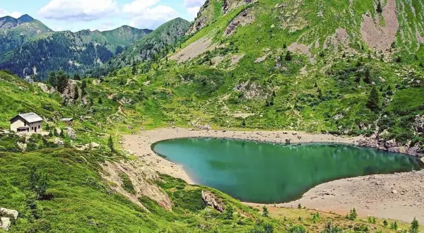 Un rifugio solitario vicino al **lago di Erdemolo**, circondato da verdi pendii montuosi e un cielo azzurro con nuvole bianche.