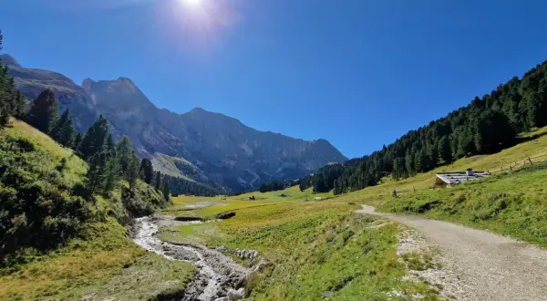 Sentiero sterrato che sale verso un rifugio alpino, con un ruscello che scorre in primo piano e montagne imponenti sullo sfondo, tipico del passo Duron.