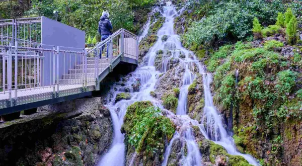 Una persona osserva le **cascate del Varone** da una passerella metallica. L'acqua scende a cascata su rocce ricoperte di muschio, immersa nel verde.