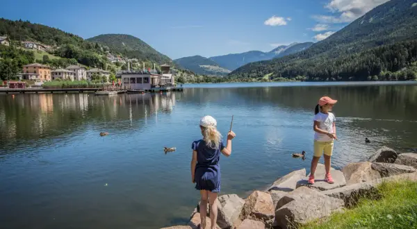 Bambine al lago di Serraia, con montagne verdi e un villaggio sullo sfondo.