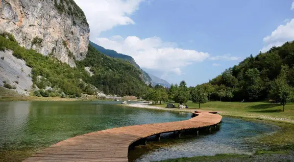 Un pontile in legno serpeggia sul lago di Andalo, circondato da montagne verdi e un cielo azzurro con nuvole bianche.