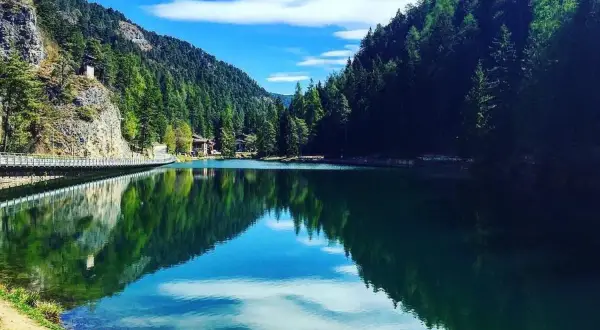 Un lago smeraldo incastonato tra montagne boscose. Una spiaggia erbosa invita al relax, mentre un ponte attraversa le acque calme.