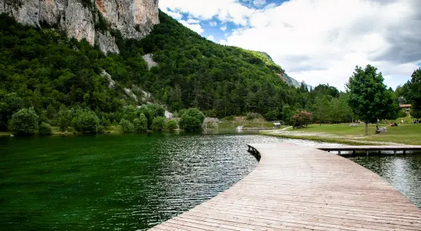 Un pontile in legno curva dolcemente sul lago di Nembia, circondato da montagne verdi e un cielo nuvoloso.