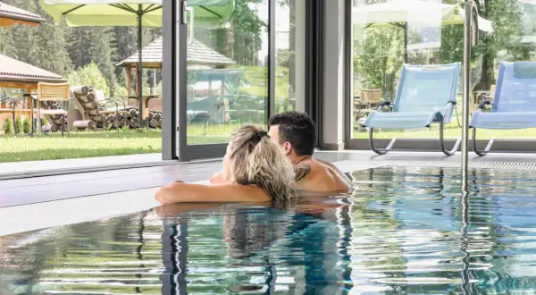 Coppia rilassata nella piscina interna di un wellness hotel Dolomia, con vista sul giardino e sulle montagne.