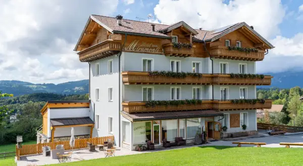 L'albergo Miramonti, con le sue balconate fiorite e il tetto in legno, si staglia contro un cielo azzurro con nuvole bianche, immerso in un paesaggio montano verdeggiante.