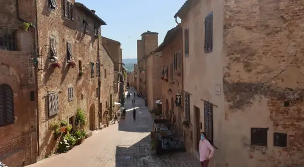 Una persona con mascherina passeggia per una stretta via acciottolata di **Certaldo Alto**, circondata da antichi edifici in mattoni con persiane in legno.