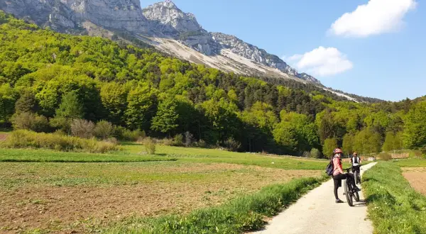 Ciclisti su un sentiero sterrato, con campi coltivati e un bosco rigoglioso ai piedi di una montagna imponente. Un passo bordala da godersi in bicicletta.