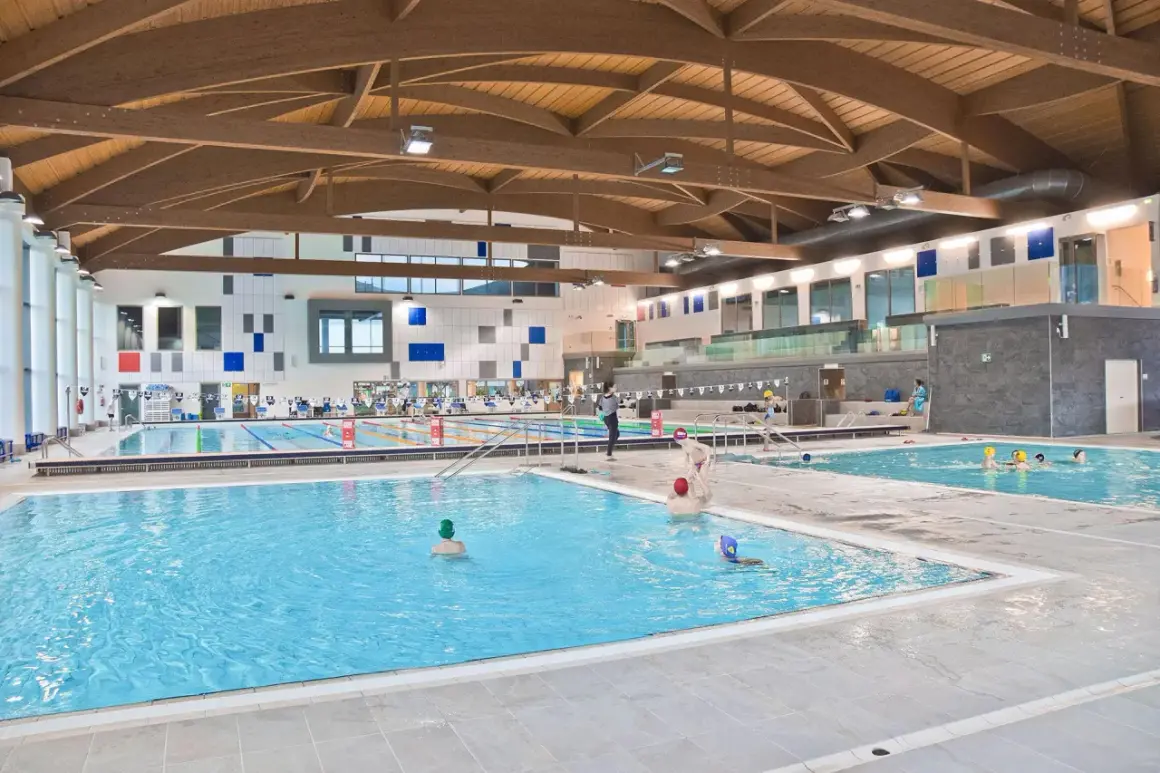 Ampia piscina coperta con tetto in legno e diverse corsie. Persone nuotano e si divertono nella piscina Pergine.