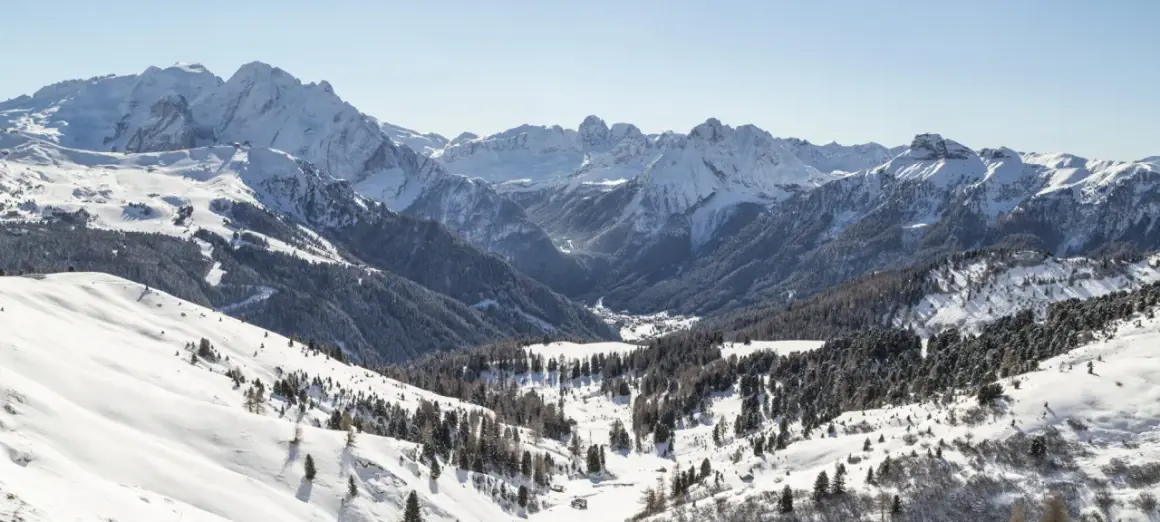 Panorami innevati di Canazei in inverno, con montagne maestose, boschi di conifere e un villaggio nascosto nella valle.