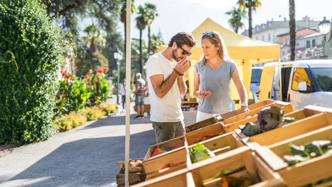 Coppia al markt Riva del Garda, annusando prodotti freschi. Verdure in cassette di legno sotto il sole.