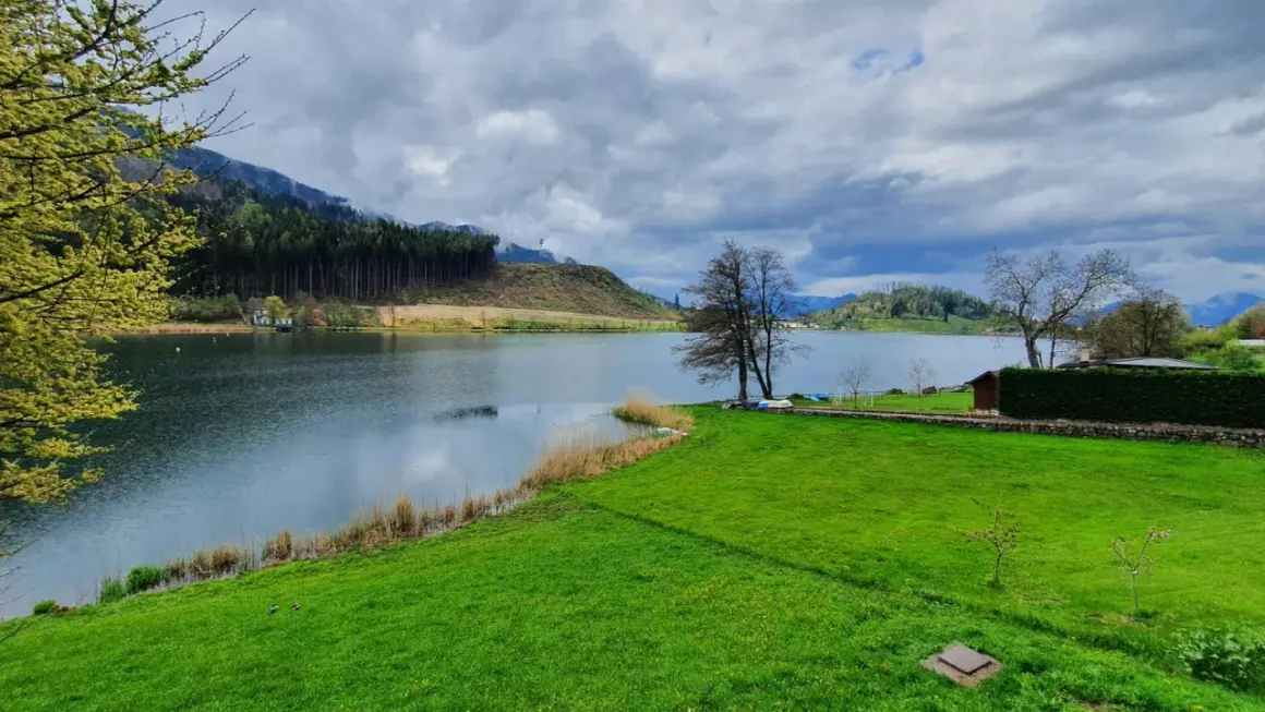 Un tranquillo lago di Pinè con verdi prati, alberi spogli e nuvole minacciose nel cielo.
