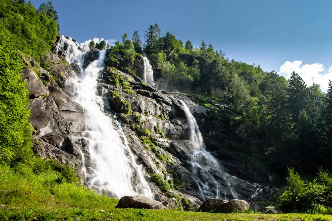Le Cascate Nardis, un imponente salto d'acqua che scende su rocce scure, circondate da un verde lussureggiante e alberi imponenti sotto un cielo azzurro.