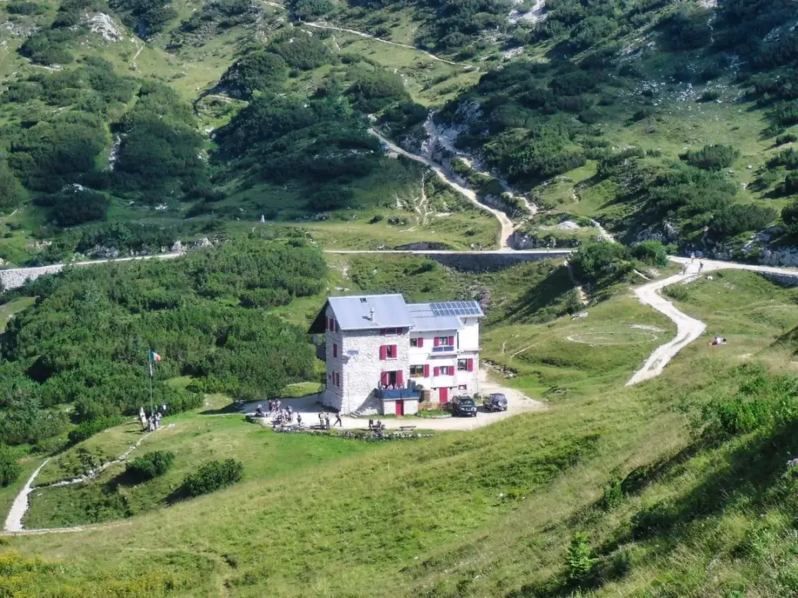 Il rifugio Scalorbi, un edificio in pietra con persiane rosse, si erge in una valle verdeggiante, circondato da sentieri e vegetazione alpina.