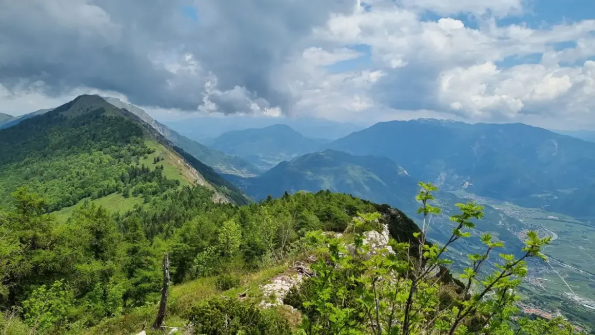 Panoramica mozzafiato dal Monte Stivo, con valli verdi e montagne azzurre sotto un cielo nuvoloso.