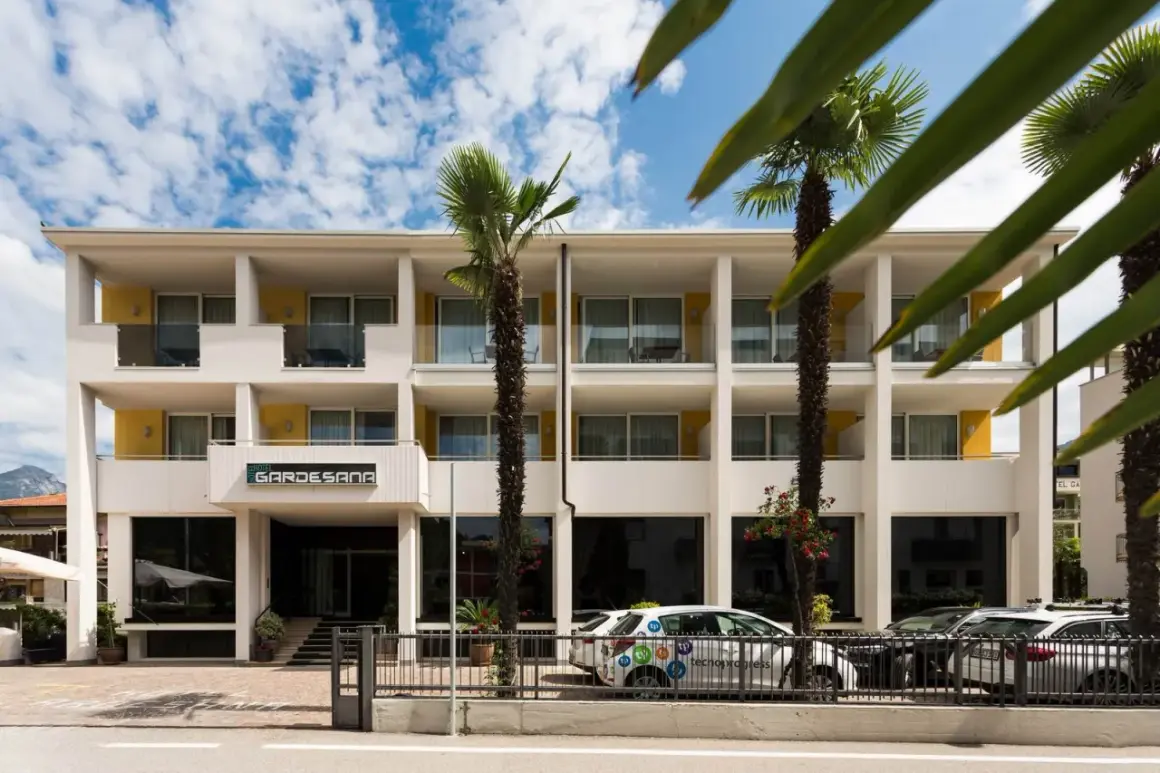 Hotel Gardesana a Riva del Garda, con palme e cielo azzurro. Un'auto bianca parcheggiata davanti.