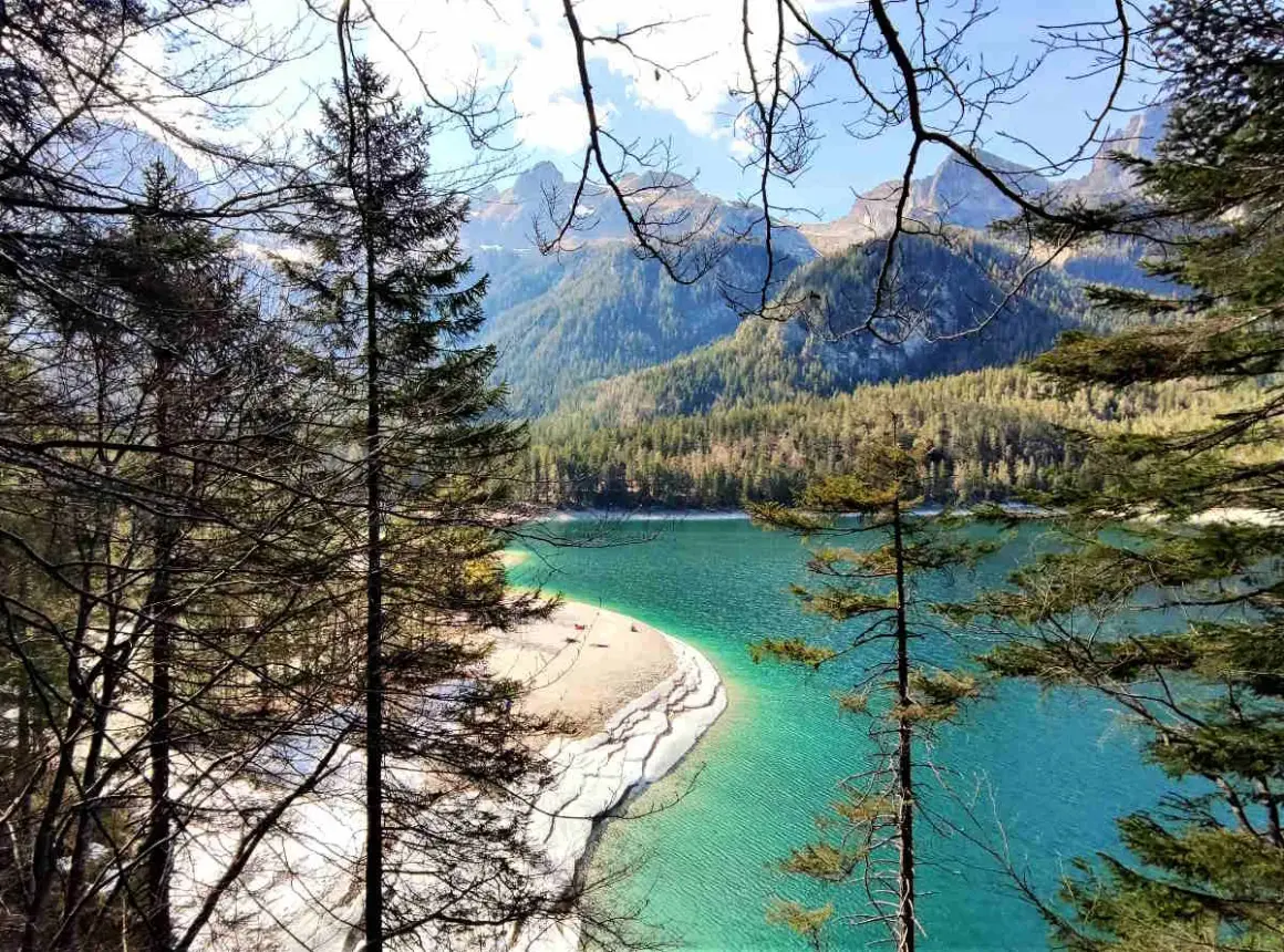Un lago turchese incorniciato da alberi e montagne nel Val di Non. La spiaggia sabbiosa invita a una sosta.