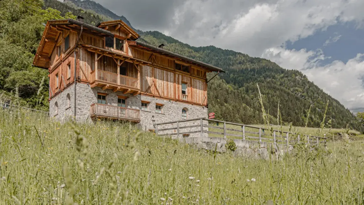 Edificio in pietra e legno con balconi, immerso nel verde alpino. Un cartello rosso indica "Hotel Rabbi".
