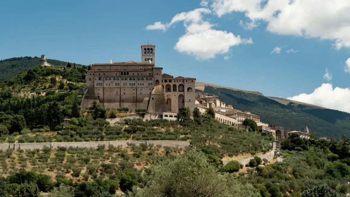 La Basilica di San Francesco domina il colle di Assisi, circondata da uliveti e montagne verdi sotto un cielo azzurro con nuvole bianche.