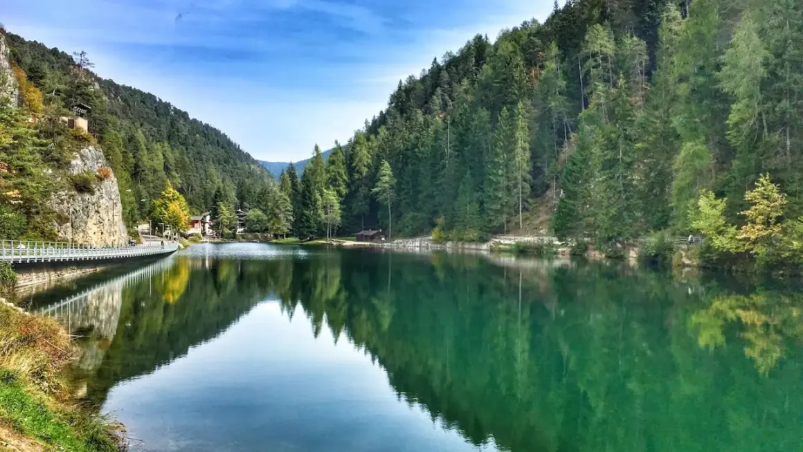 Un **lago smeraldo** incastonato tra montagne boscose, con un ponte pedonale che riflette le sue acque cristalline.