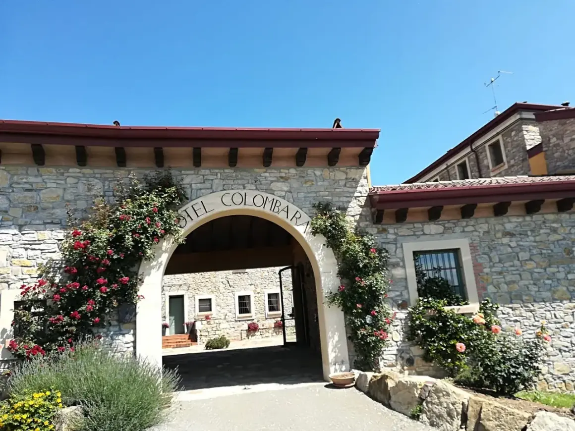 Ingresso in pietra del relais La Colombara, con rose rosse rampicanti e un arco che porta a un cortile interno.