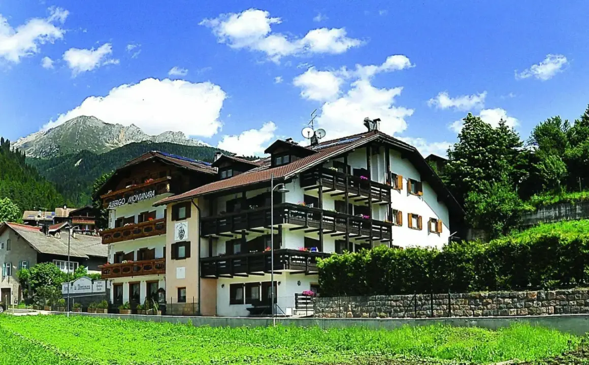 Albergo La Montanara a Predazzo, con le sue balconate in legno e le montagne sullo sfondo, incorniciato da un cielo azzurro e nuvole bianche.
