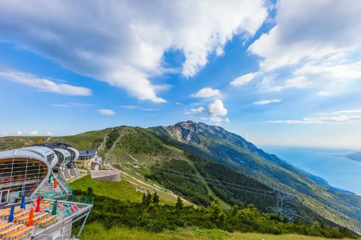 Edificio moderno con terrazza panoramica sul Monte Baldo, con vista sul lago e montagne verdi sotto un cielo azzurro con nuvole bianche.