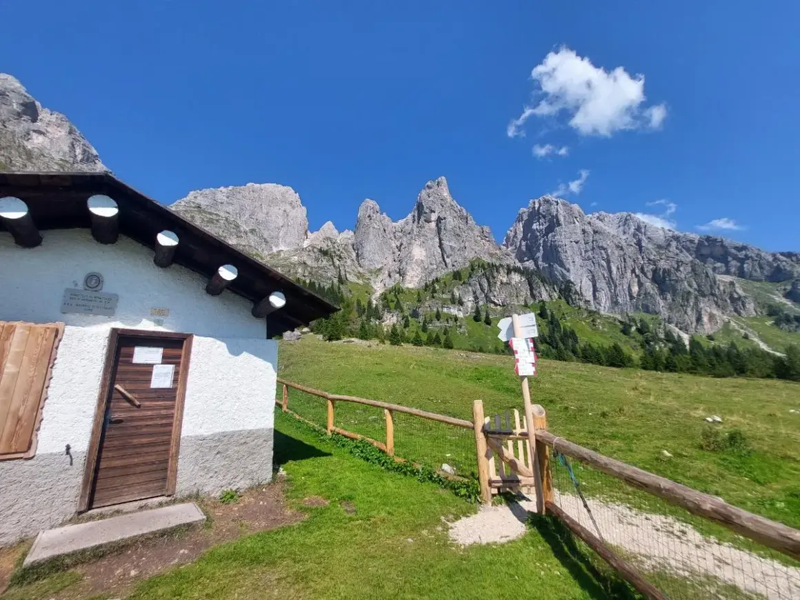 Malga Cavallera: sentiero con cartelli direzionali e rifugio bianco ai piedi delle maestose montagne.