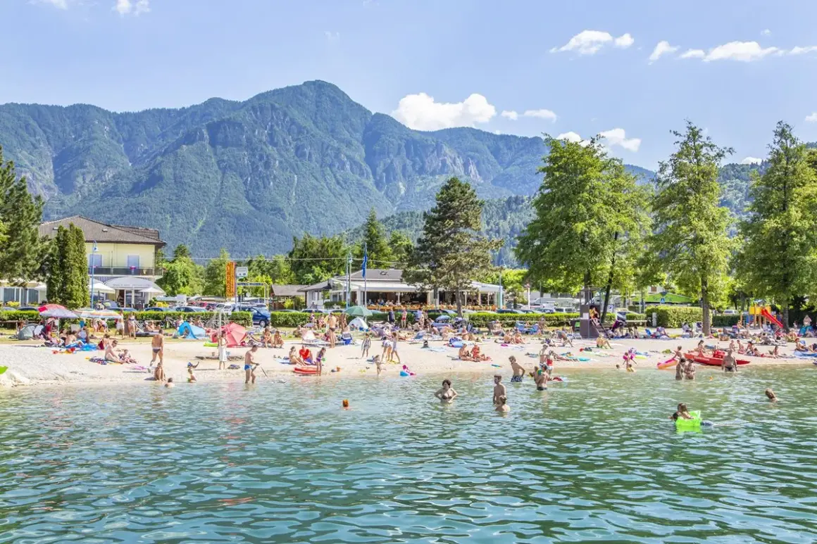 Spiaggia affollata al camping Riviera Caldonazzo, con gente che prende il sole e nuota nel lago, montagne sullo sfondo.