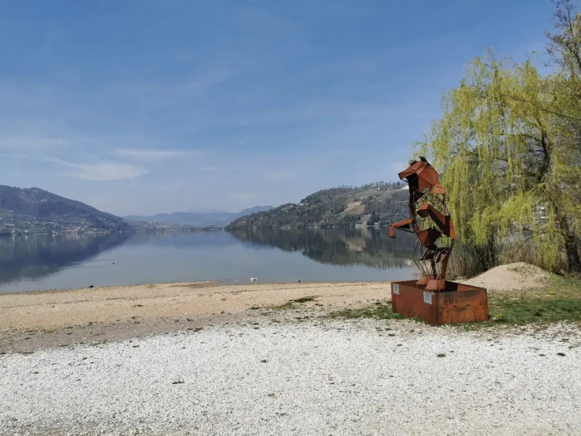 Scultura metallica di un cane sulla riva del lago di Caldonazzo, con montagne riflesse sull'acqua calma.