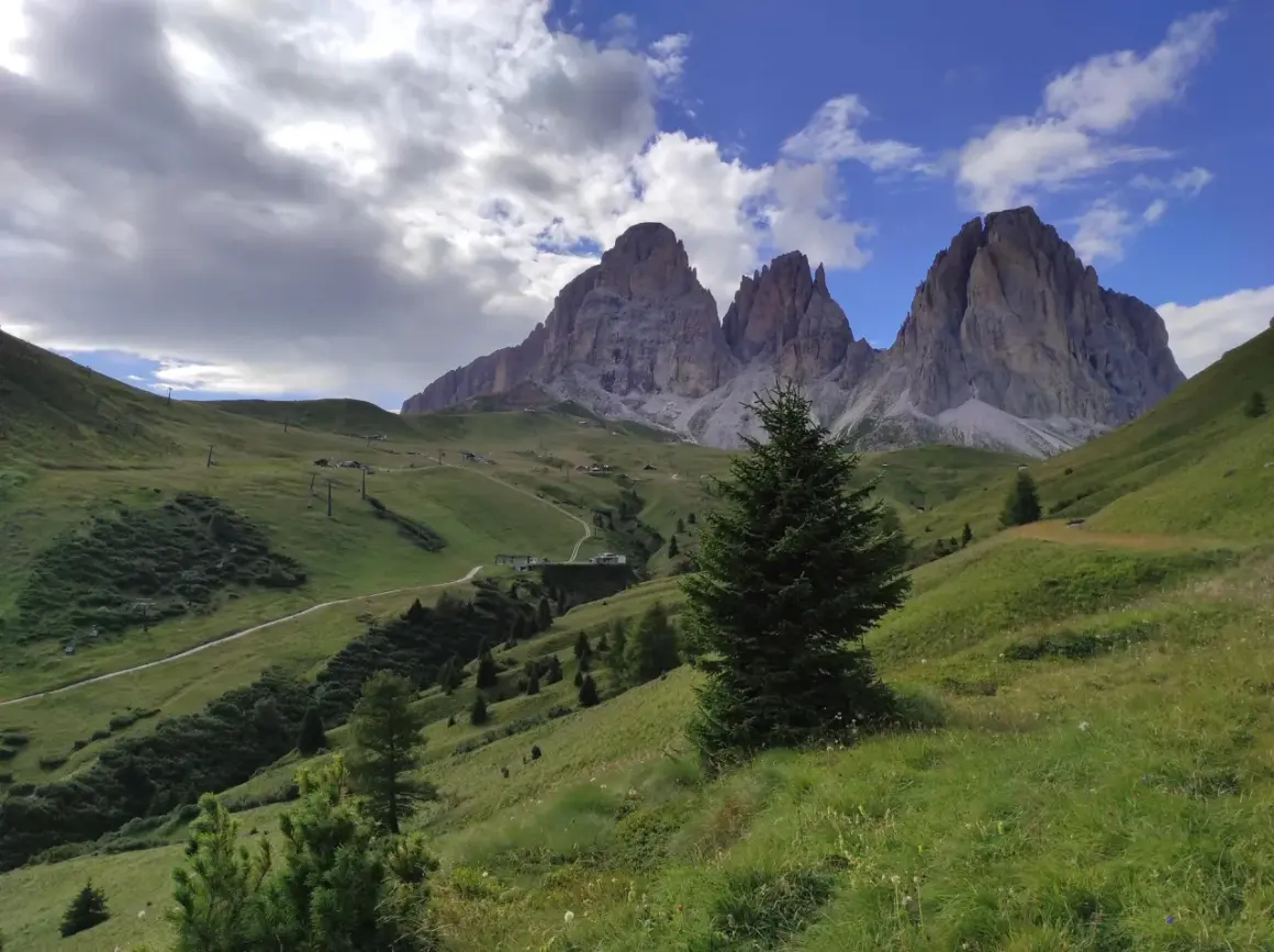 Verdi pendii alpini verso le imponenti cime del Sassolungo, con sentieri e impianti di risalita. Immagina le cascate basaltiche di Canazei in lontananza.