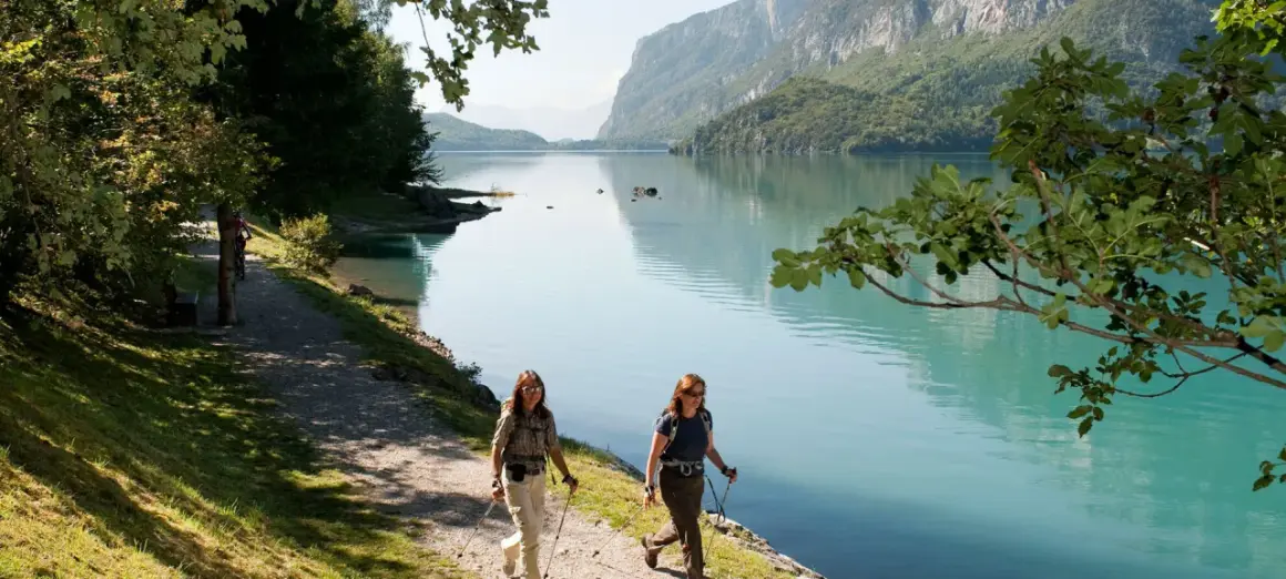 Due escursioniste camminano lungo il sentiero del lago di Molveno in estate, con montagne verdi sullo sfondo.