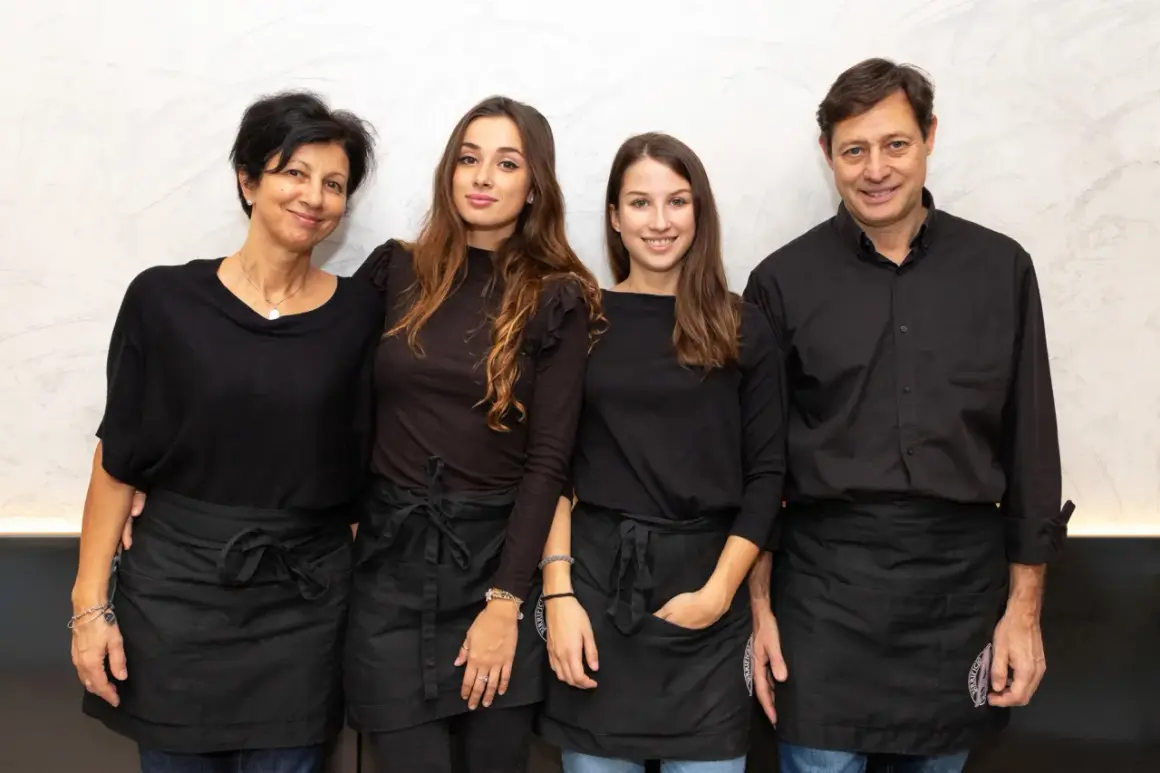 Four people, two women and two men, wearing black shirts and aprons, stand together smiling.