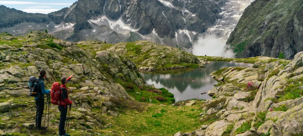 Due escursionisti ammirano un lago alpino e montagne imponenti, seguendo il sentiero della pace.